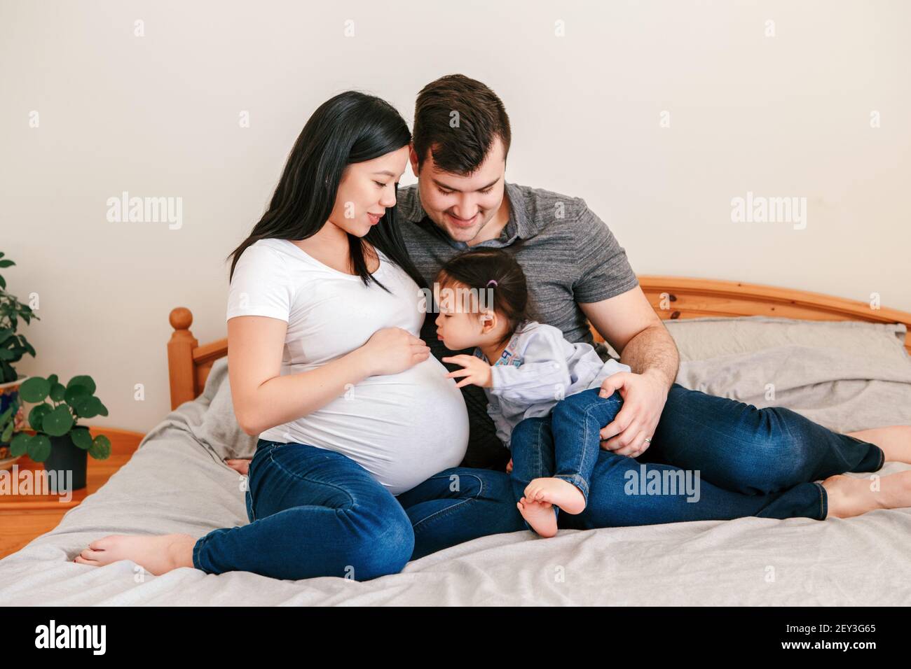 Famiglia Asiatica Cinese incinta donna e uomo caucasico con bambina seduta  sul letto a casa. Madre, padre e bambina in attesa Foto stock - Alamy