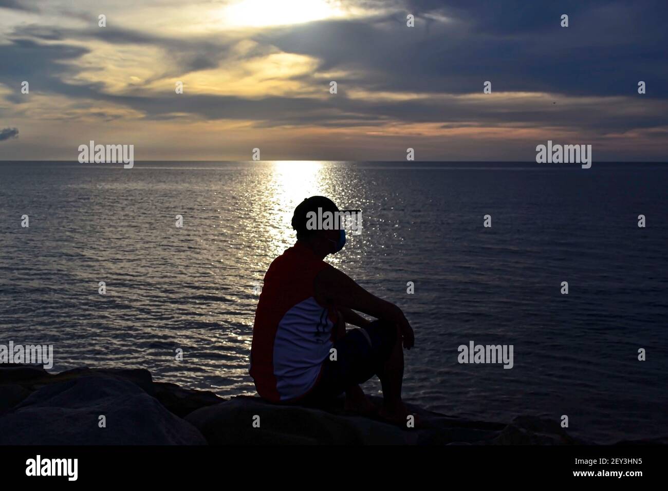 Uomo seduto su una roccia che guarda l'alba in mare Foto Stock