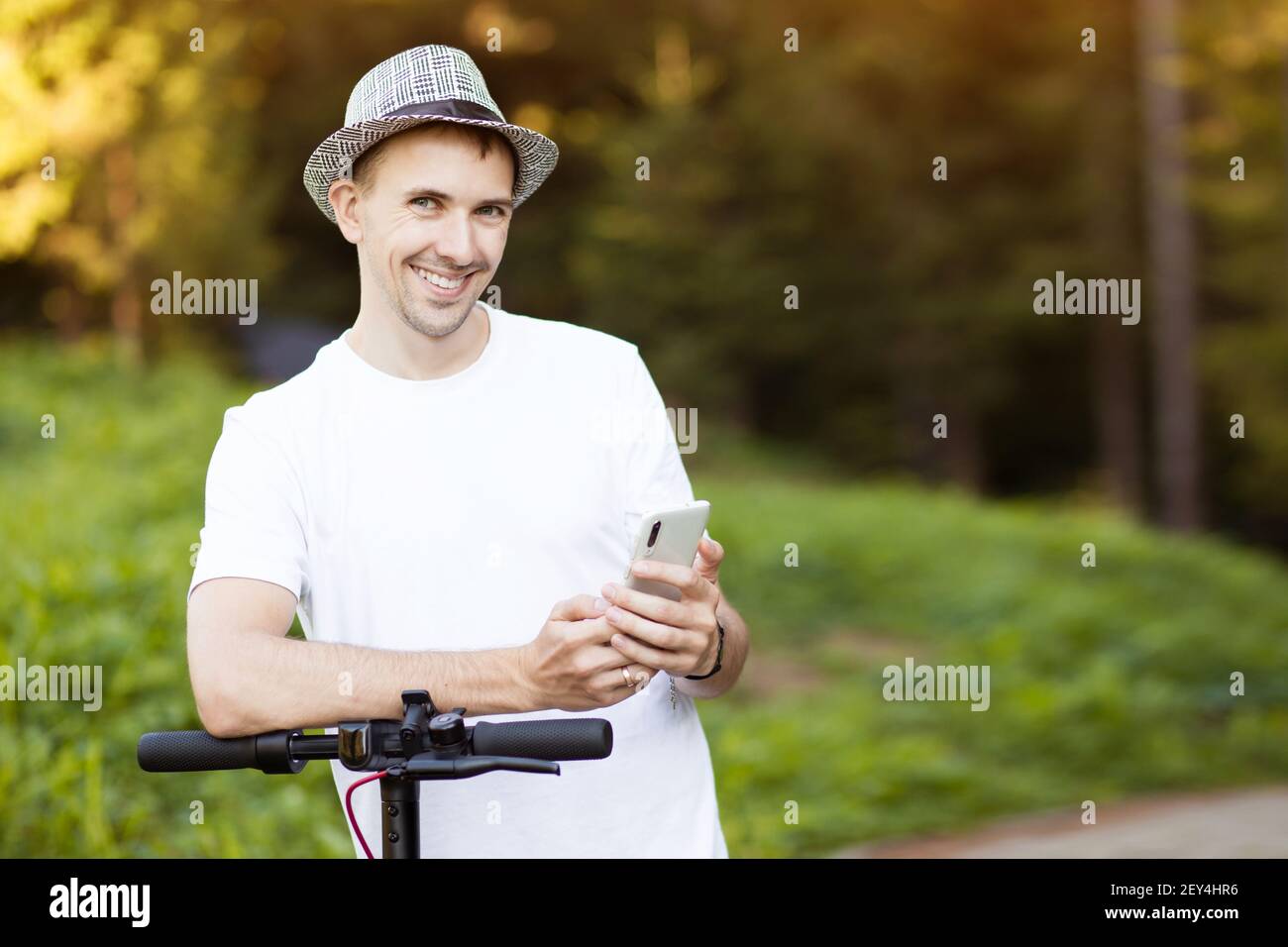 Giovane uomo in cappello usa il telefono cellulare, prenota online noleggio scooter elettrico in città. Trasporti innovativi Foto Stock