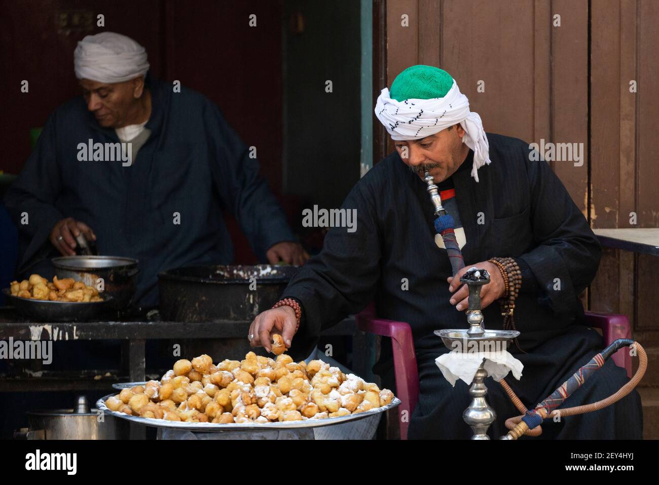 Ritratto di un uomo egiziano locale che fuma un tubo di narghilè e che mangia dolci fritti in un caffè (ahwa) nel quartiere islamico del Cairo, Egitto Foto Stock