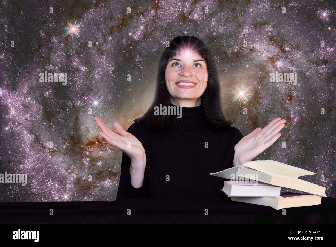 Una ragazza positiva in vestiti neri si siede ad un tavolo con i libri sullo sfondo del cielo stellato. Sulla fronte della ragazza è presente un luminoso simbolo stellare Foto Stock