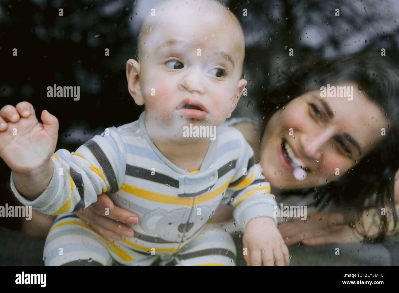 Bambino divertente e mummia alla finestra piovosa durante la quarantena Foto Stock