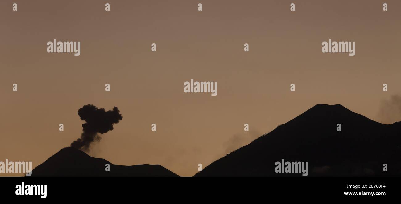 Il vulcano Fuego (fuoco) dalle silhouette emette un soffio di cenere accanto al vulcano Acatenango al crepuscolo vicino Antigua, Guatemala Foto Stock