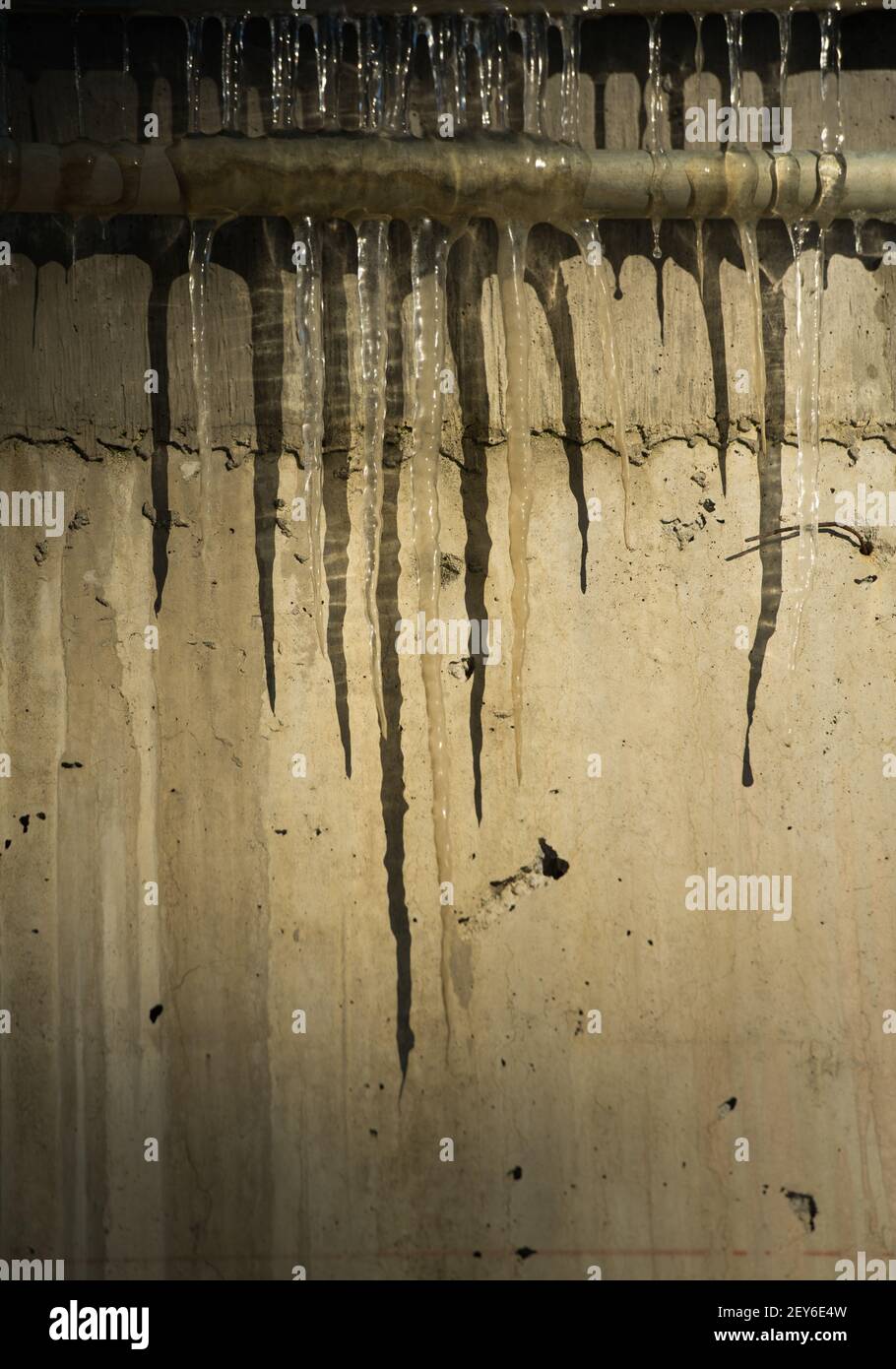 ghiaccioli congelati trasparenti e appuntiti appesi alle ombre della fusione industriale di tubi su cemento grungy esterno parete di fondo verticale Foto Stock