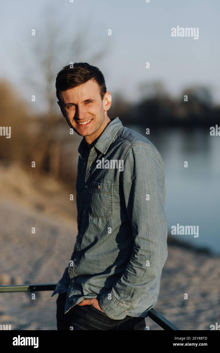 Uomo sorridente in camicia jeans in piedi fuori con le mani in tasca. Ritratto dell'uomo Foto Stock