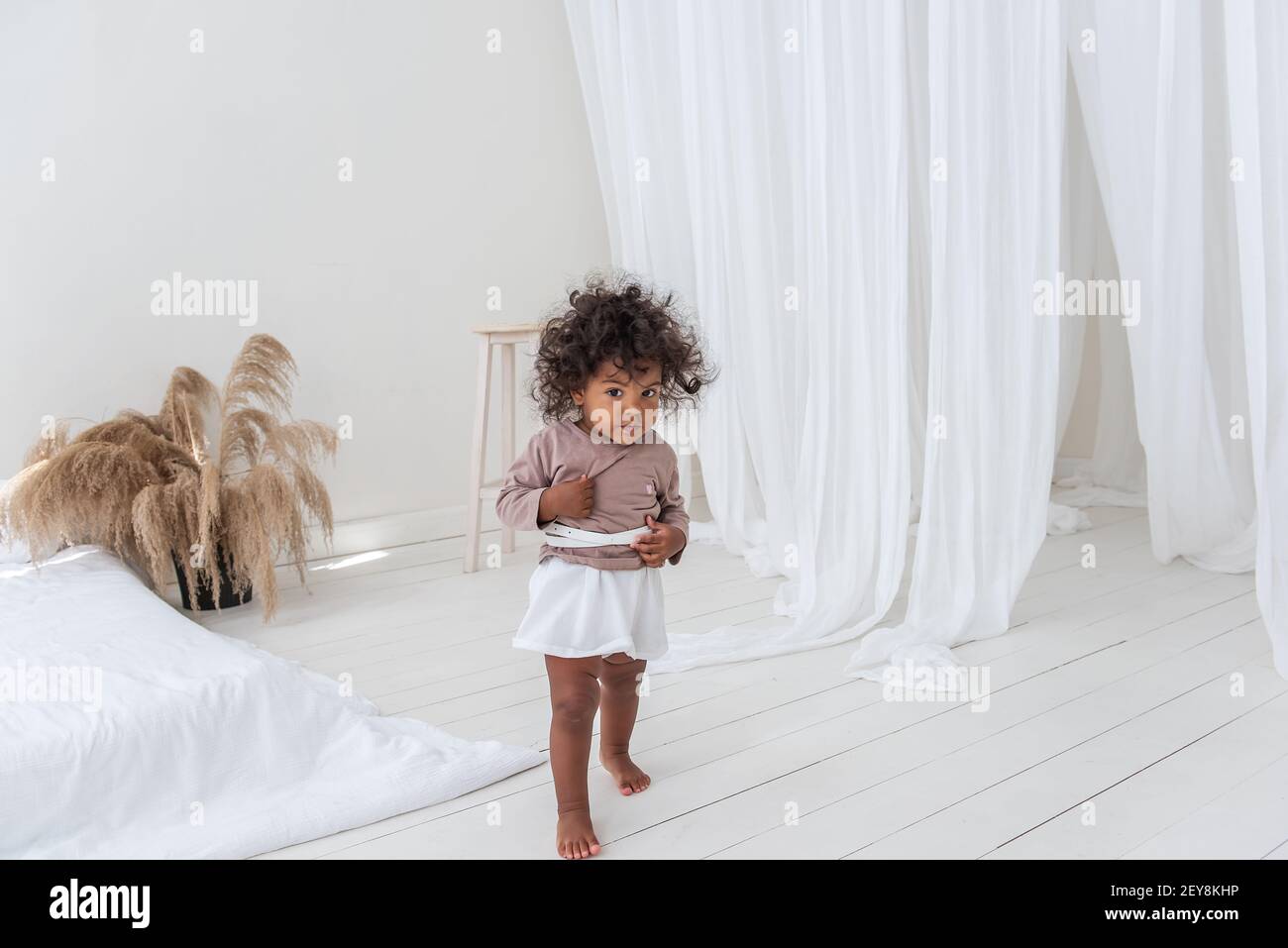 Piccola ragazza afroamericana riccia-capelli gioca, corre dal letto bianco, ha divertimento, ride, sorrisi. Il concetto di adottare una famiglia multietnica. Scandina Foto Stock