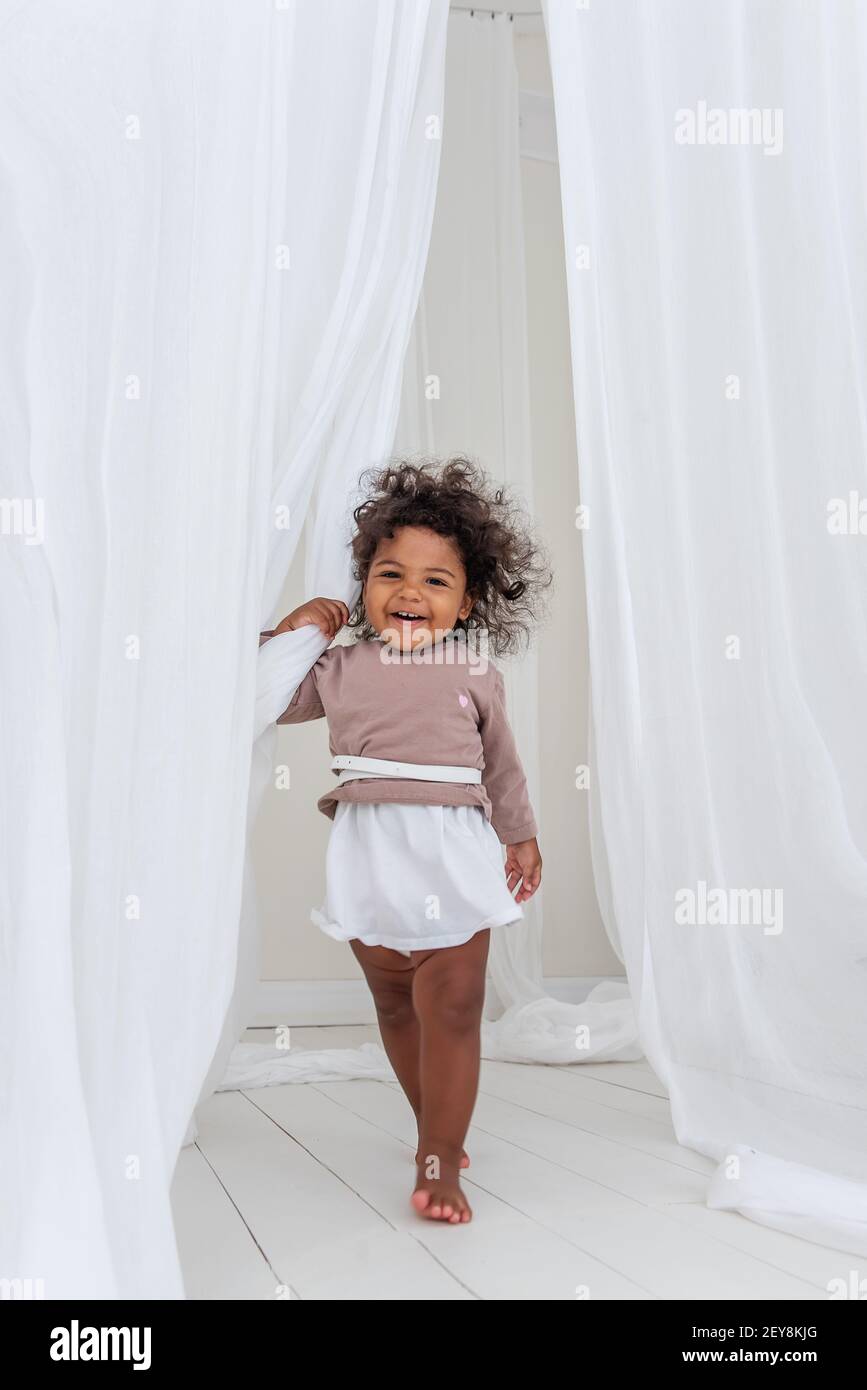 La ragazza americana africana poco curly ha danze divertenti di risata fra il tessuto bianco arioso delle tulle. Interni minimalisti, stile scandinavo. Felice infanzia. Il Foto Stock