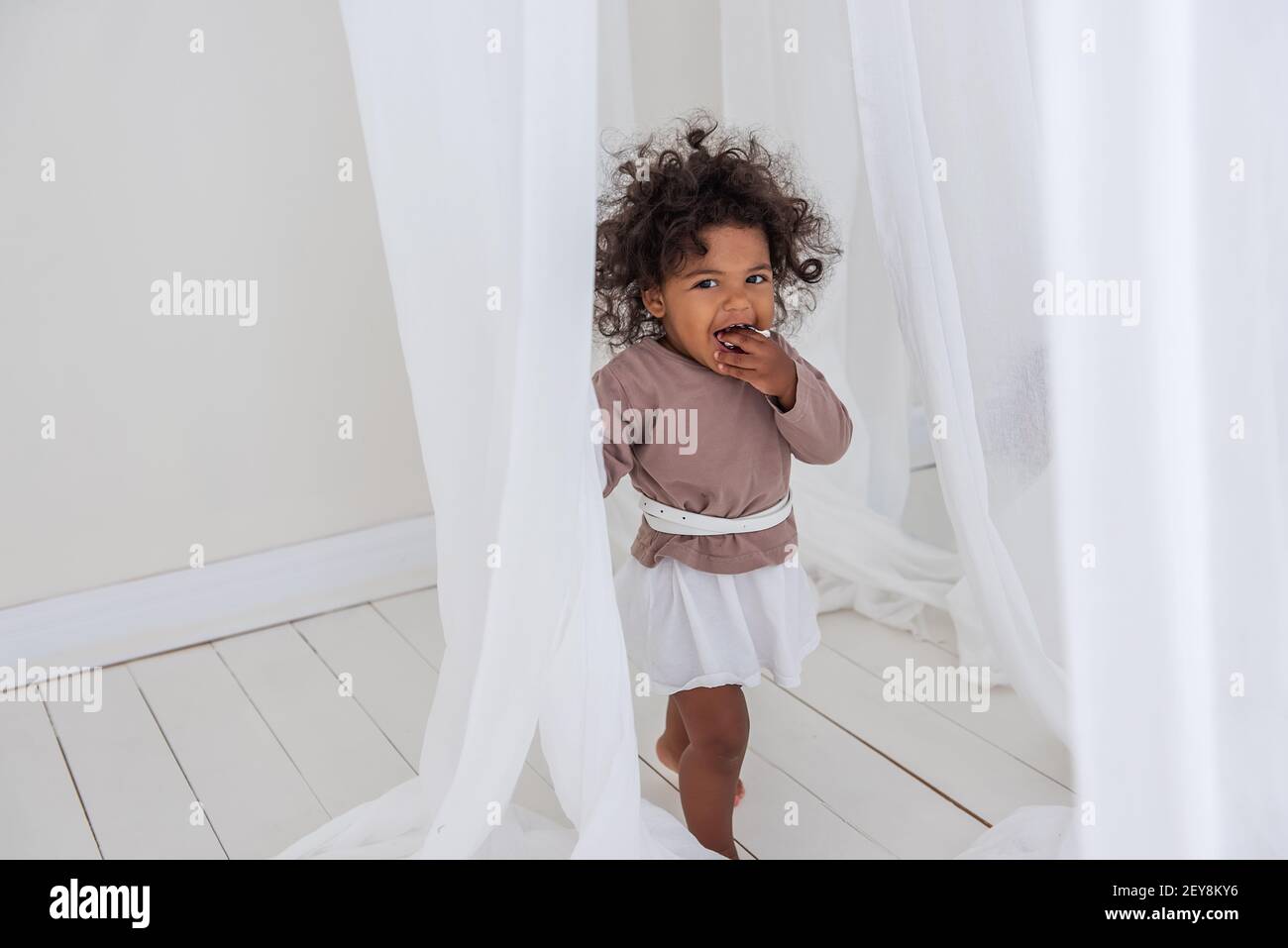 La ragazza americana africana poco curly ha danze divertenti di risata fra il tessuto bianco arioso delle tulle. Interni minimalisti, stile scandinavo. Felice infanzia. Il Foto Stock