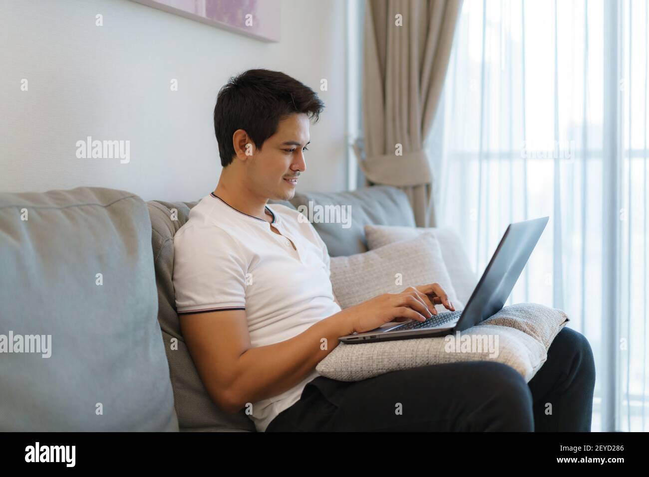Un uomo asiatico che lavora a casa, chattando online con un collega sul suo laptop mentre si trova sul divano in soggiorno a casa. Foto Stock