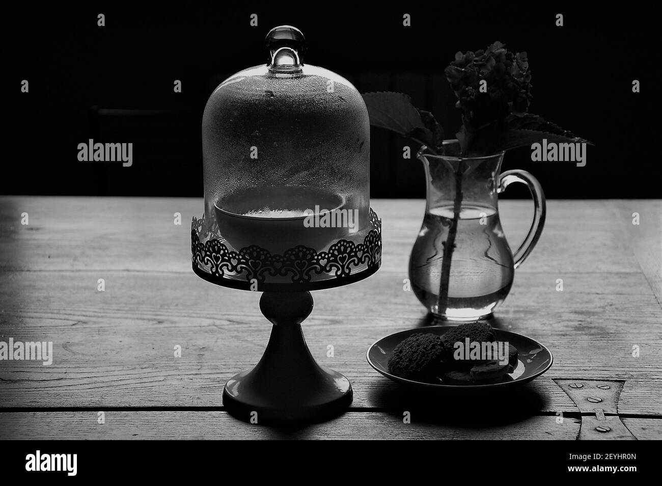 bianco e nero ancora vita di un cake stand, bevanda calda e un vaso di vetro di fiori sul tavolo wodden in cucina al coperto Foto Stock