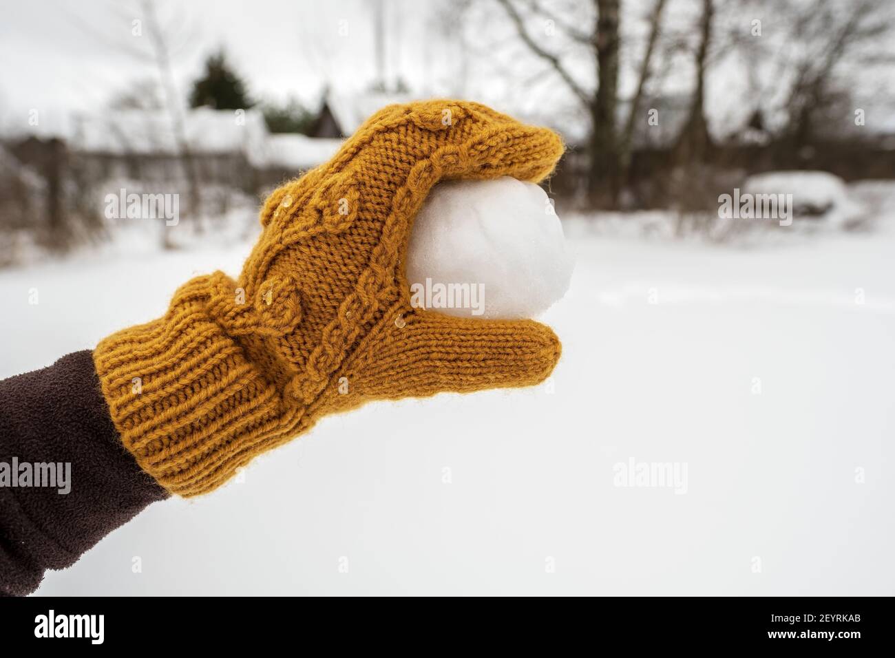 Una mano in un mitten giallo tiene una palla di neve, nel cortile sullo sfondo di un orto innevato. Giochi attivi invernali e divertimento. Foto Stock