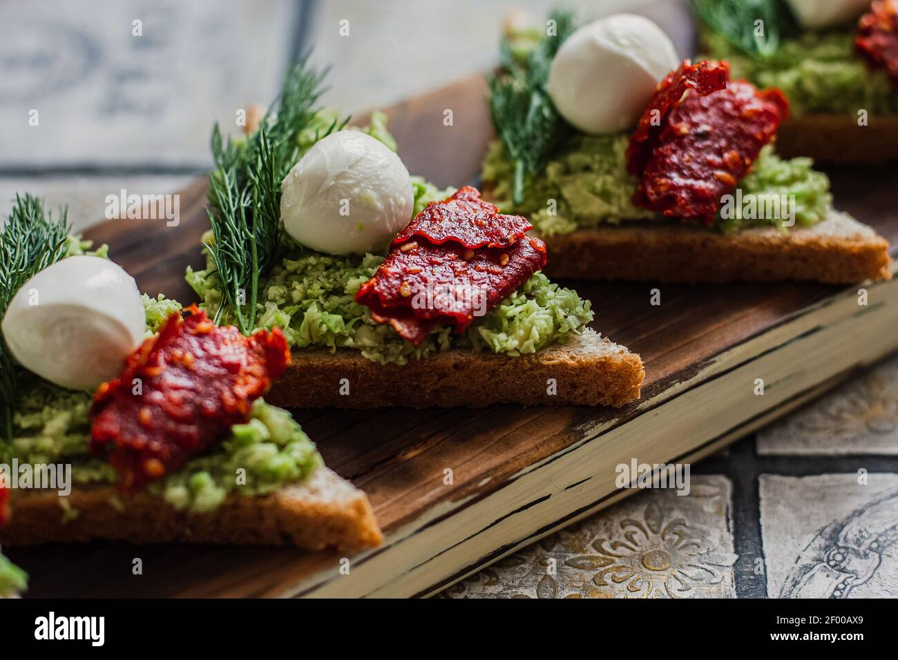 Mini crostini triangolari bruschetta con avocado, mozzarella e tomatos essiccati sulla tavola rustica sullo sfondo piastrellato d'annata Foto Stock