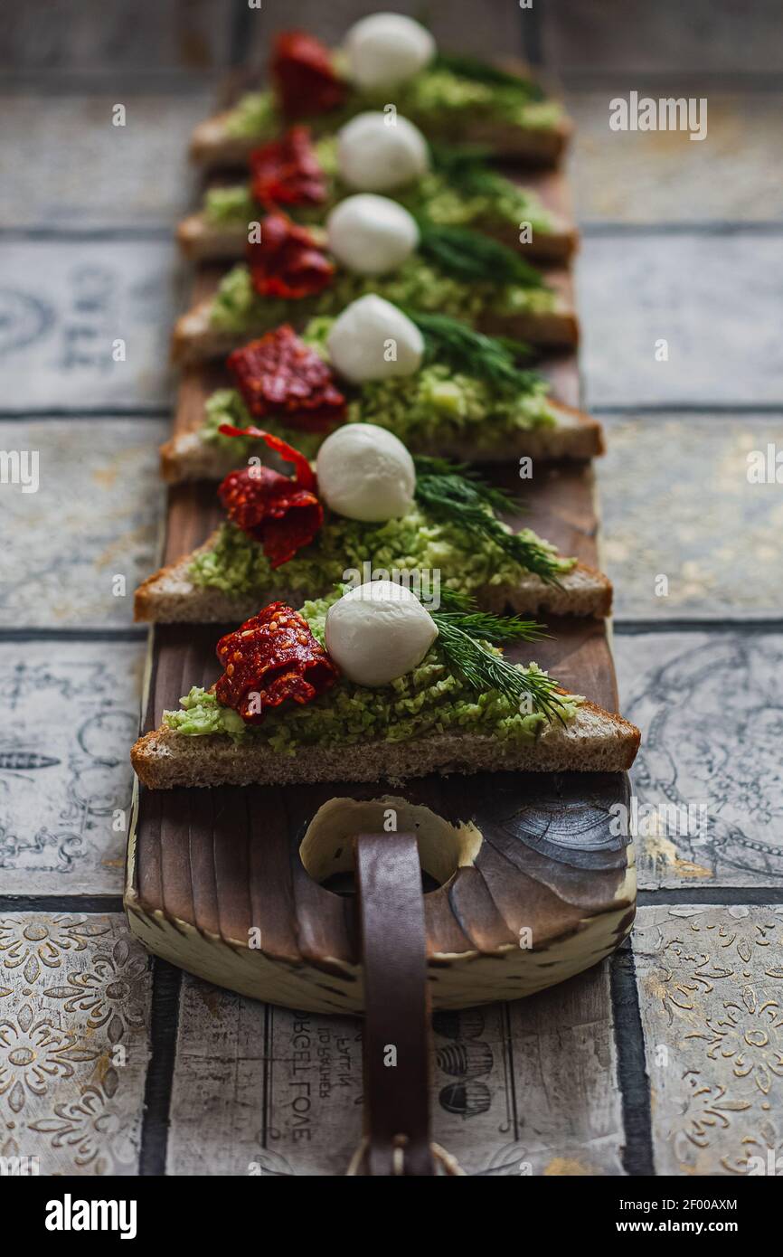 Mini crostini triangolari bruschetta con avocado, mozzarella e tomatos essiccati sulla tavola rustica sullo sfondo piastrellato d'annata Foto Stock