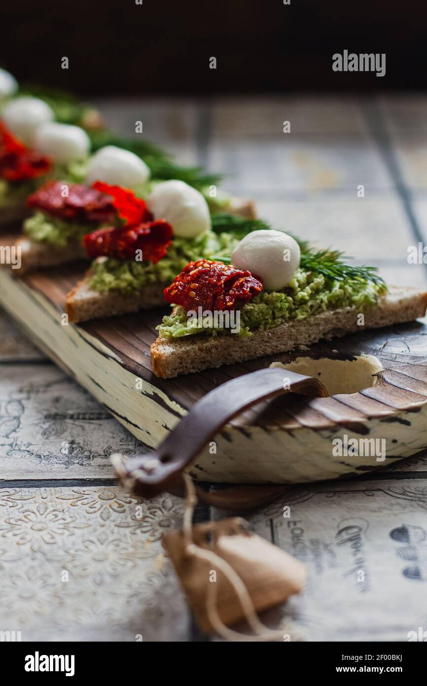 Mini crostini triangolari bruschetta con avocado, mozzarella e tomatos essiccati sulla tavola rustica sullo sfondo piastrellato d'annata Foto Stock