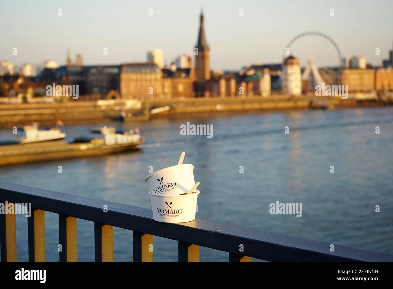 Due tazze di carta di yogurt ghiacciata "Yomaro" sulla ringhiera del ponte Oberkasseler sul fiume Reno con la storica città vecchia sullo sfondo sfocato. Foto Stock