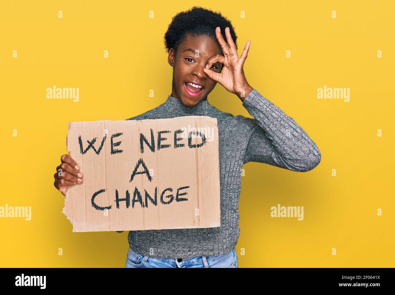 Giovane ragazza afro-americana che tiene abbiamo bisogno di un banner di cambiamento sorridente felice fare ok segno con mano su occhio guardando attraverso le dita Foto Stock