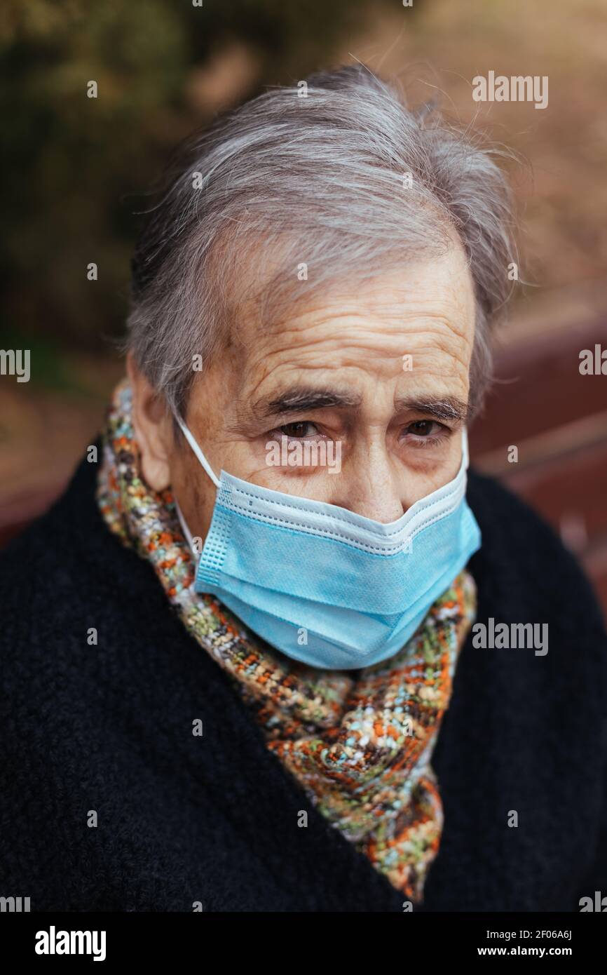 Ritratto di donna anziana con maschera facciale guardando la macchina fotografica mentre si siede su una panca del parco Foto Stock
