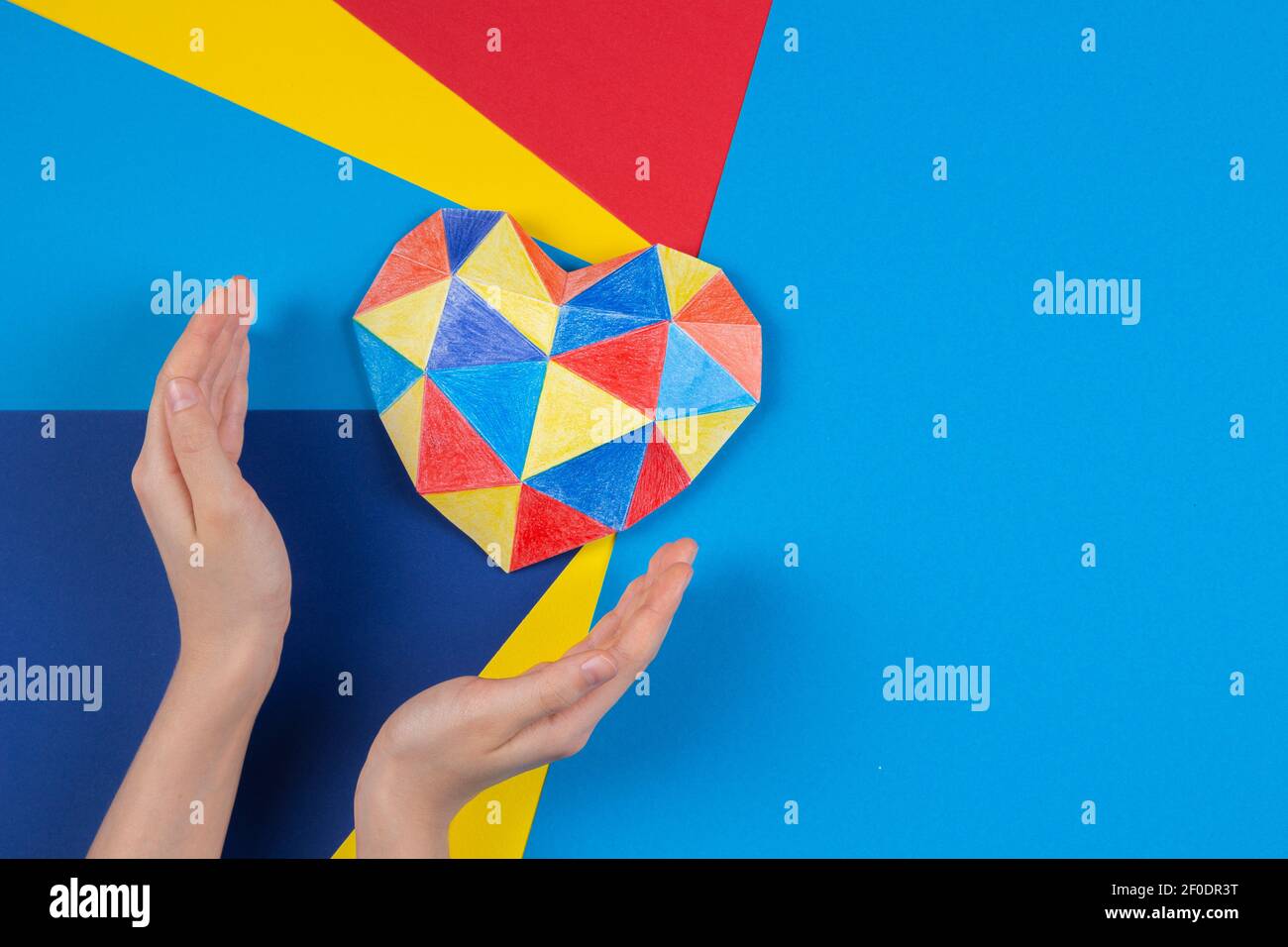 Mani del bambino con cuore colorato di carta su sfondo di carta multicolore. Concetto di giorno di consapevolezza autismo mondiale Foto Stock