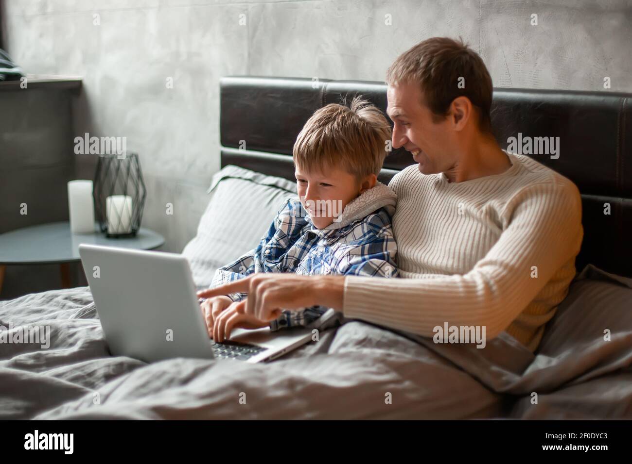 Fine settimana a casa. Papà e figlio si trovano a letto sotto una coperta e giocano sul computer portatile. Festa di Bachelor. Padre e figlio sono alla ricerca di un dono per la loro madre Foto Stock