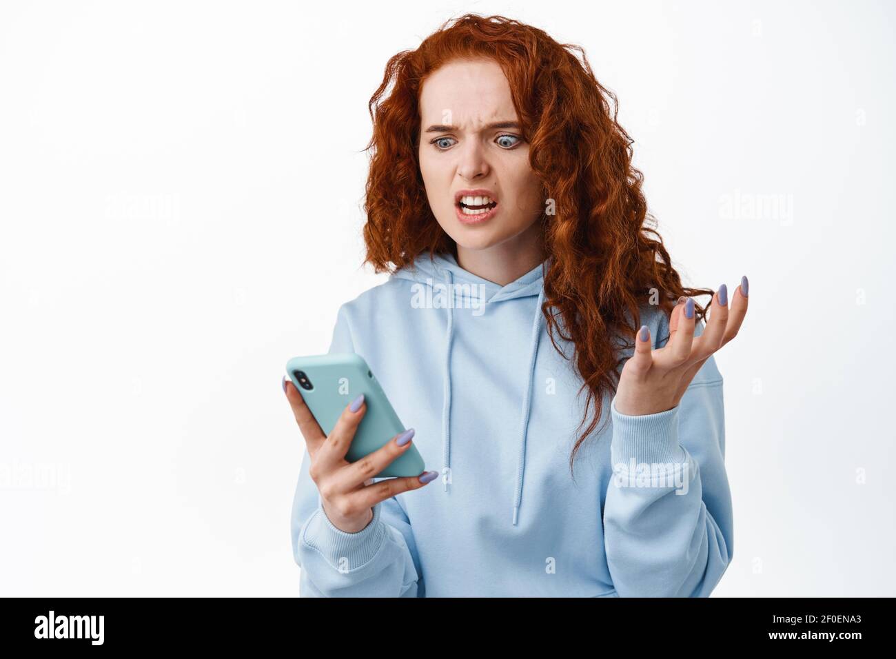 Arrabbiata e infastidita donna rossa grimacing pazza, fissando allo schermo dello smartphone, leggendo il messaggio fastidioso, in piedi disturbato su sfondo bianco Foto Stock