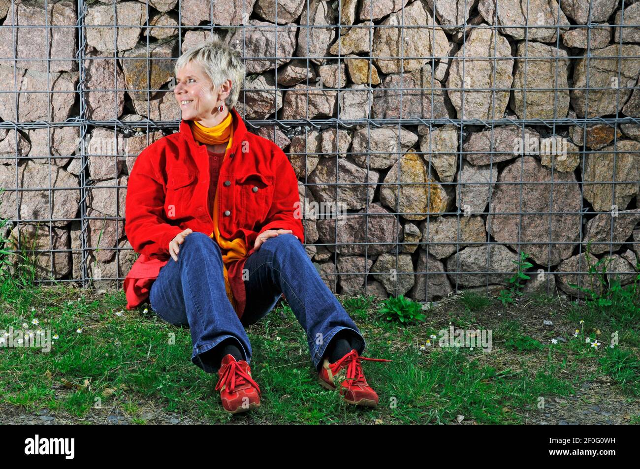 Ritratto a corpo pieno di una donna matura in jeans blu, giacca rossa, scarpe sportive rosse e sciarpa gialla seduta sul pavimento con la schiena contro un baratto Foto Stock