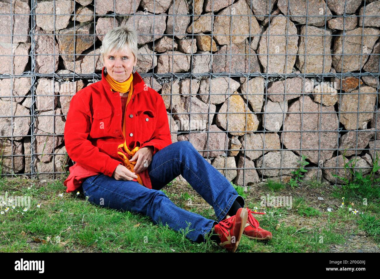 Ritratto a corpo pieno di una donna matura in jeans blu, giacca rossa, scarpe sportive rosse e sciarpa gialla seduta sul pavimento con la schiena contro un baratto Foto Stock