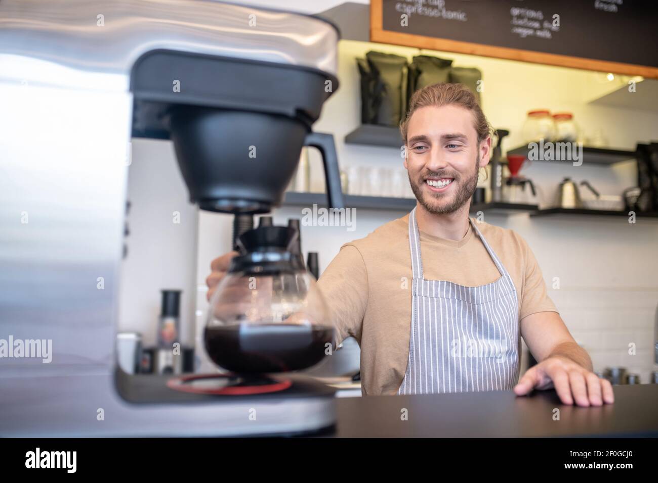 Uomo sorridente con caffettiera accanto alla macchina per il caffè dietro il bancone Foto Stock