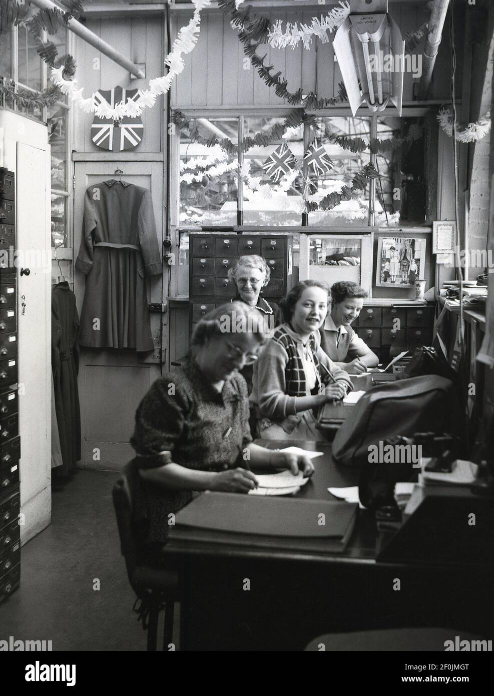1953, storico personale femminile che lavora in un piccolo ufficio presso l'azienda di abbigliamento Hepworths, Leeds, Inghilterra, UK. I martinetti di Bunting e Union sono sul muro, con una foto della regina Elisabetta II e del principe Filippo - indossando un kilt - con due ragazzini. I conigli e le bandiere celebrano l'incoronazione della Regina al trono britannico. La Providence Works su Clay Pit Lane era la casa di Joseph Hepworth e Son, una compagnia di abbigliamento iniziata nel 1865 e che nel 1905 aveva 143 negozi. Più tardi nel secolo, Hepworths divenne Next. Foto Stock