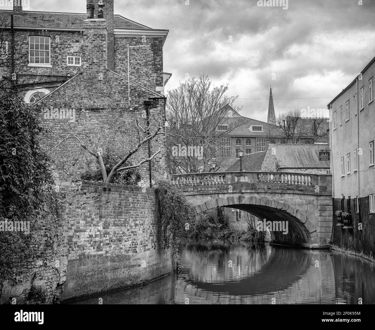 Un vecchio ponte attraversa un fiume tra due edifici. Il ponte si riflette nell'acqua e una guglia della chiesa si può vedere in lontananza. Foto Stock