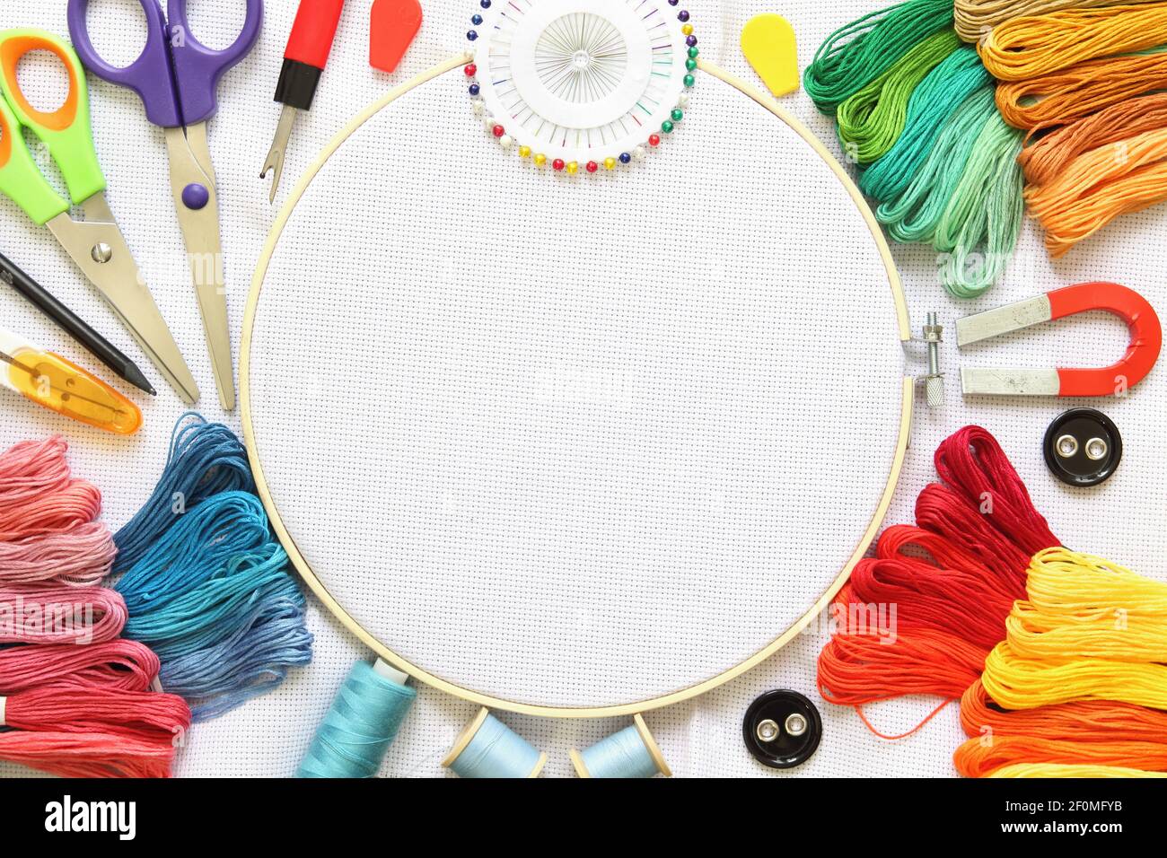 Cerchio da ricamo e accessori multicolore su tela bianca con rocchetti di filo, ago e forbici. Foto Stock