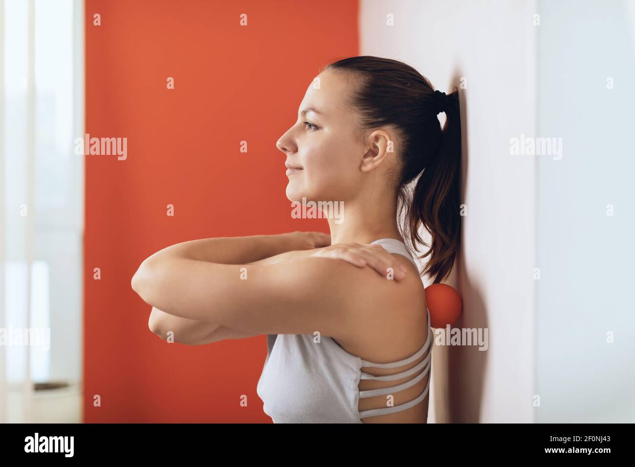 Bella donna esegue il rilascio miofasciale dei muscoli iper-flessibili della schiena con una palla di massaggio vicino alla parete. Il concetto di affaticamento della schiena preve Foto Stock