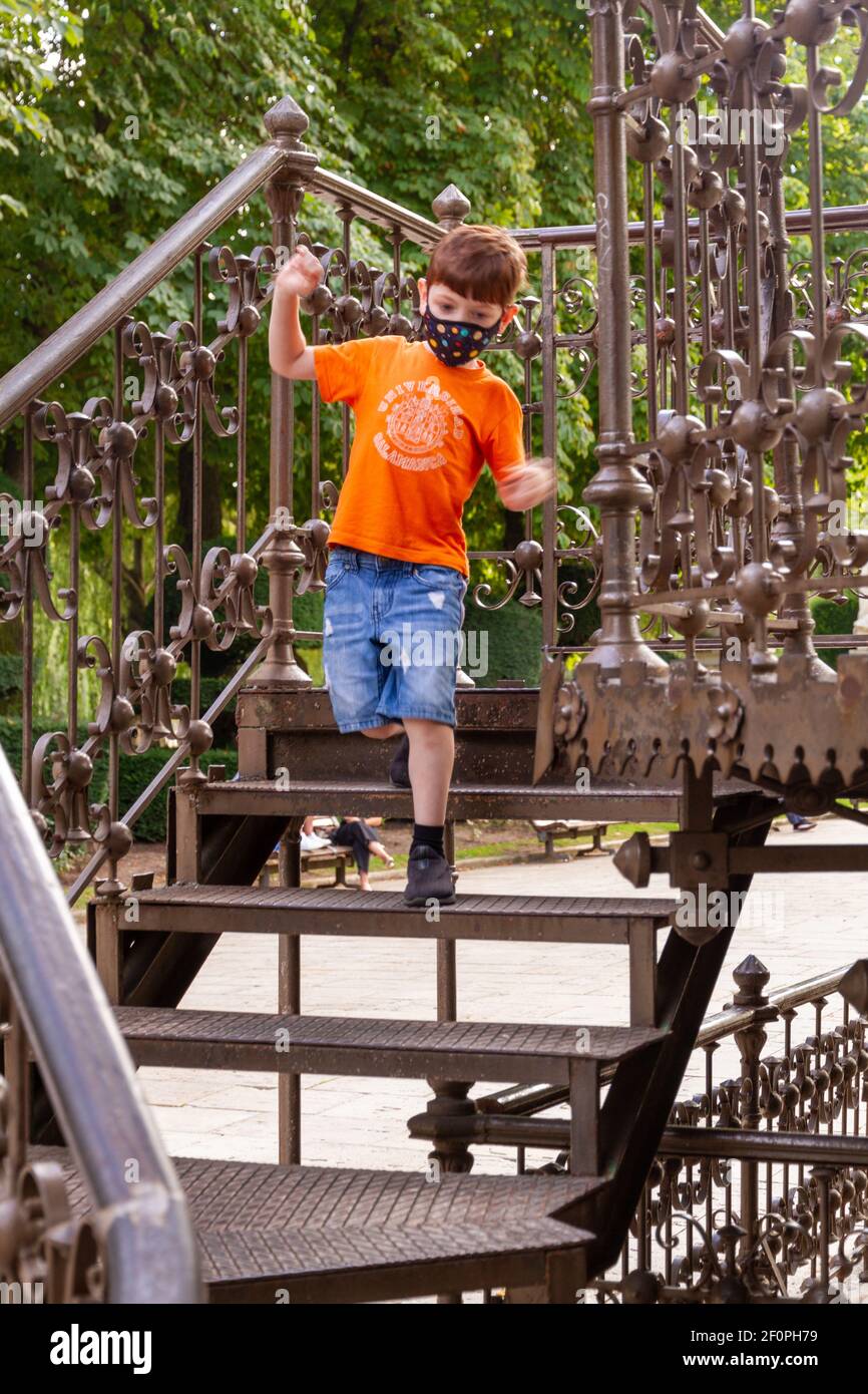 Un ragazzo carino rosso che indossa una maschera facciale e un t-shirt arancione che cammina sulle scale di un chiosco metallico una giornata di sole Foto Stock