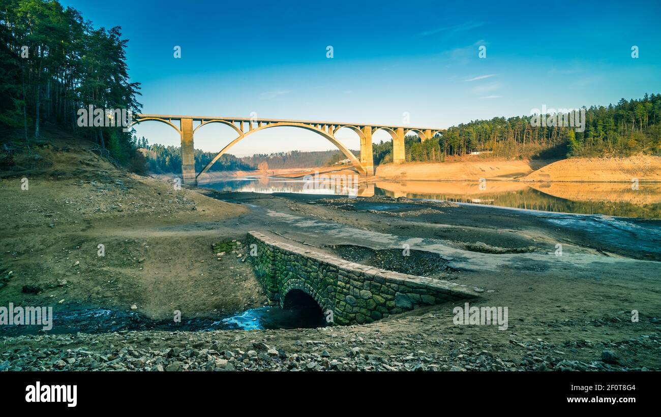 Alto viadotto ad arco attraverso la diga vuota di Orlik e l'originale letto del fiume Moldava. Contrasto di grande cemento e piccolo ponte di pietra vecchia strada. Podolsko, Repubblica Ceca. Foto Stock