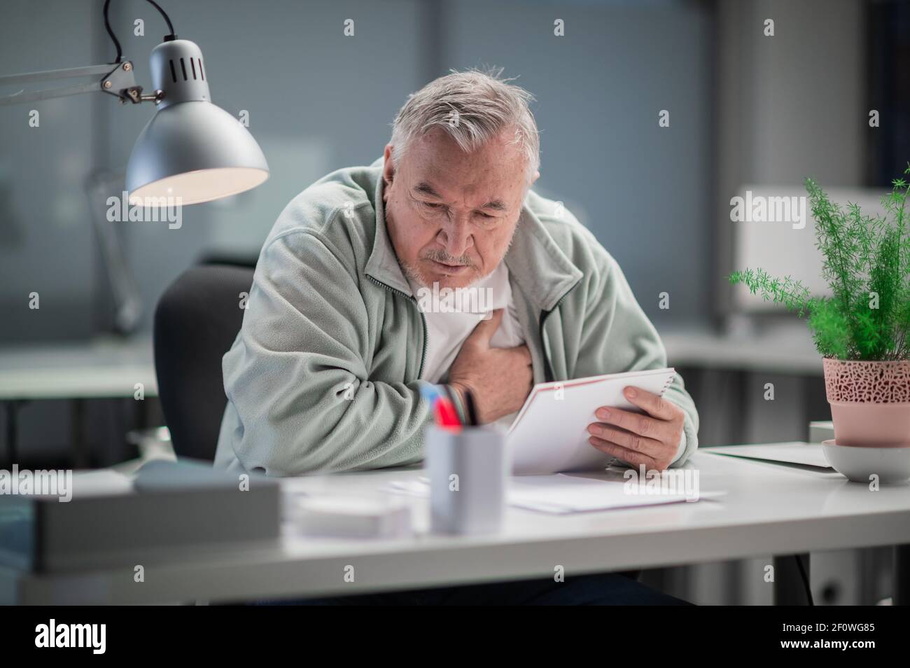 Nel suo ufficio, un uomo dai capelli grigi sentì un dolore nel suo cuore mentre lavorava, si siede con la mano sul petto Foto Stock