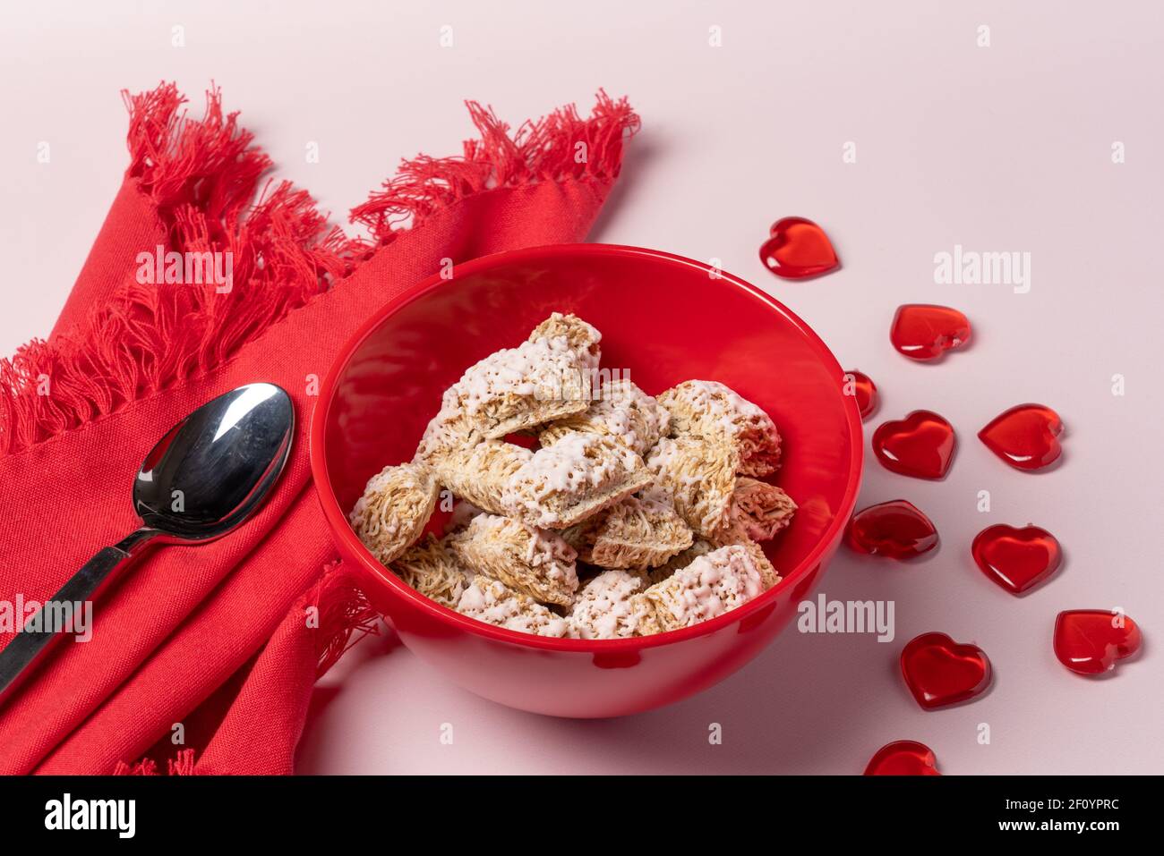 colazione romantica o valentina con ciotola rossa di cereali di grano ghiacciati, tovagliolo e cucchiaio con tavola di cuori rossi sparso su sfondo rosa chiaro Foto Stock