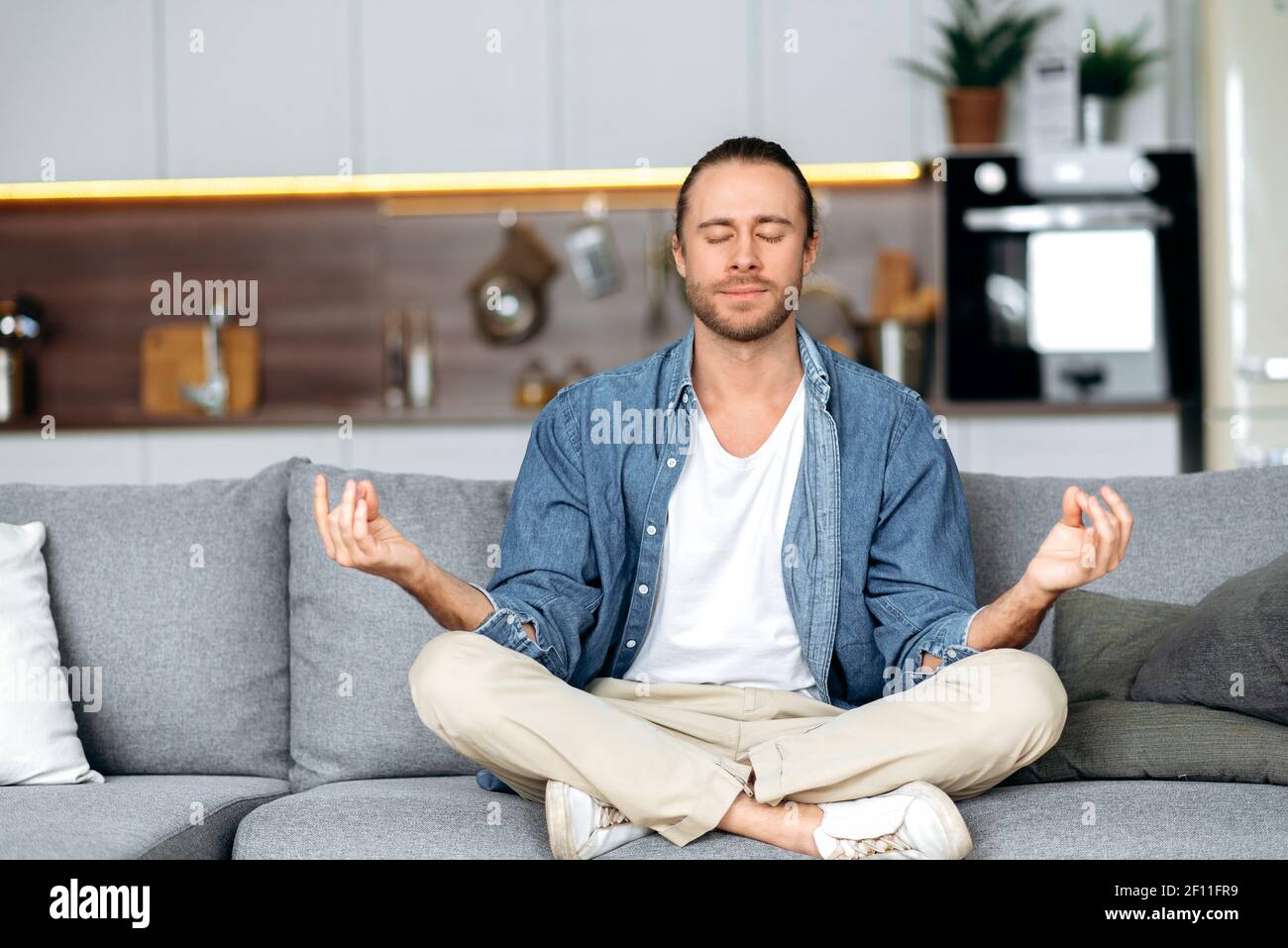 Calmo e spensierato ragazzo caucasico vestito in un elegante abbigliamento casual medita mentre si siede in posizione lotus sul divano a casa con gli occhi chiusi, rilassante. Concetto di calma e armonia Foto Stock