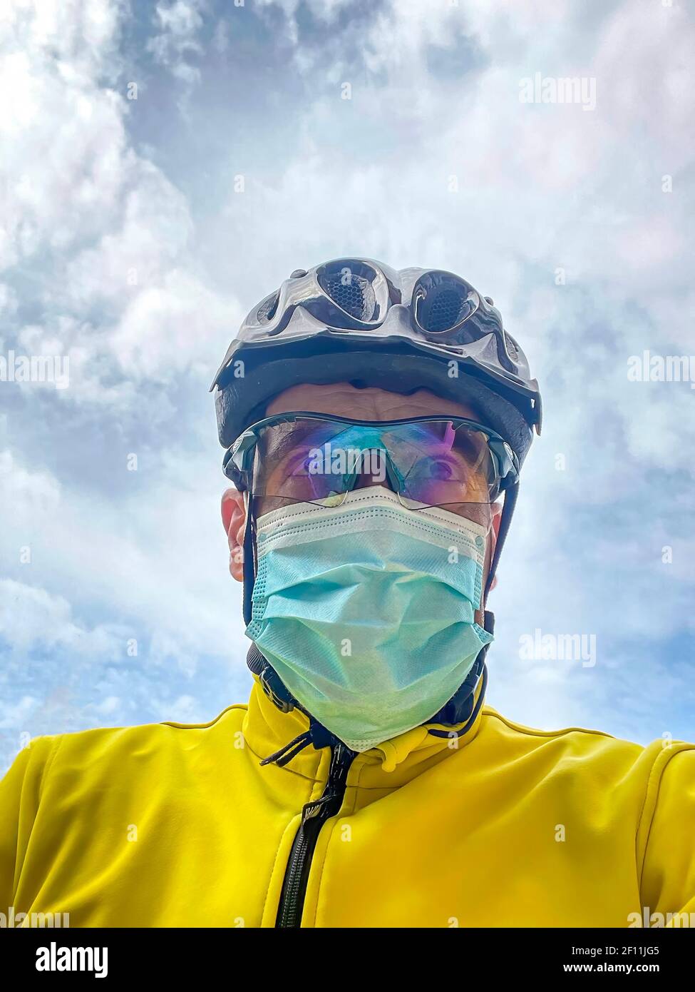 ritratto di un ciclista con casco, occhiali e giacca gialla, in bicicletta con maschera, ritratto verticale contro un cielo nuvoloso Foto Stock
