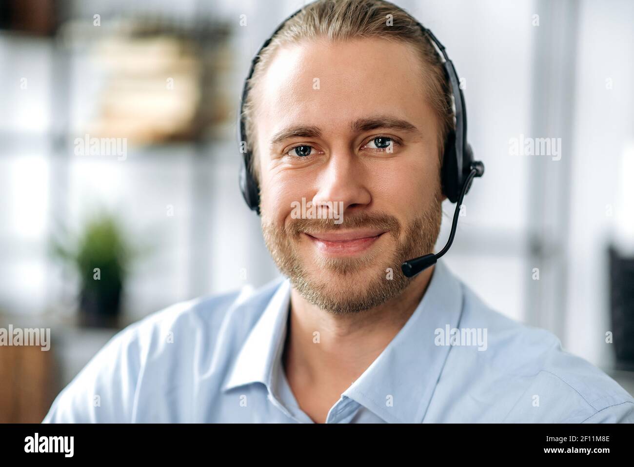 Ritratto in primo piano di un operatore soddisfatto e attraente del call center o del leader aziendale. Uomo d'affari caucasico in cuffia e camicia formale guarda la fotocamera e amichevole sorridente Foto Stock