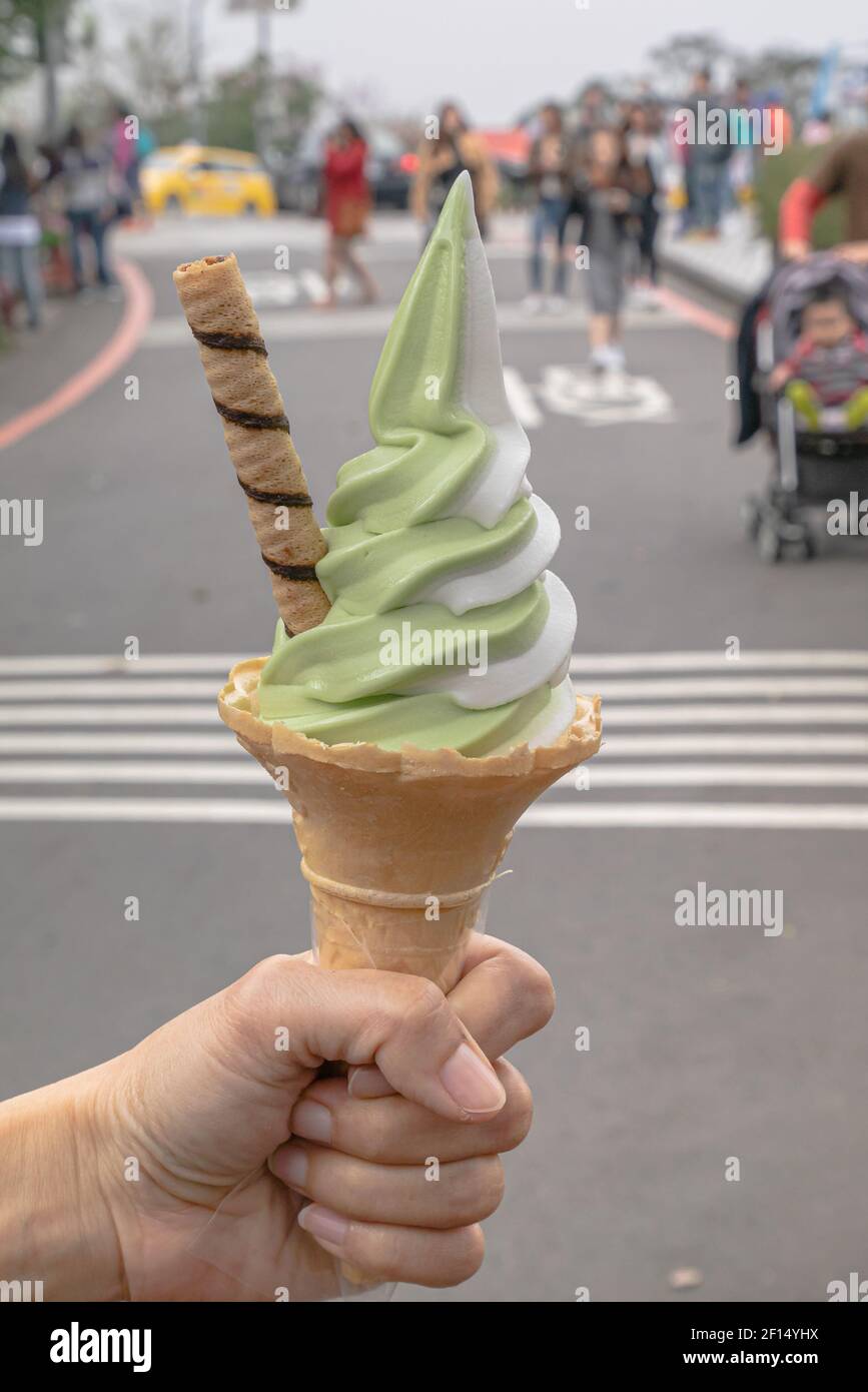 Cono gelato, un primo piano di tè verde matcha e vaniglia soft-servito con cono di waffle in mano tenendo in viaggio strada con gente sfondo a Tai Foto Stock