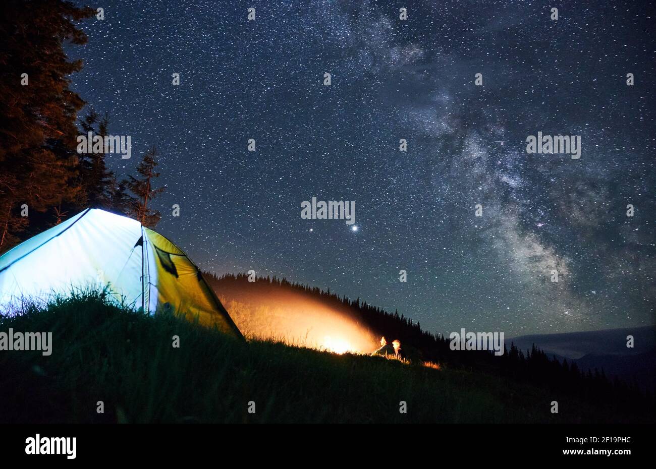 Due persone seduti accanto al falò nel loro campeggio con tenda illuminata e di guardare la fiamma. Notte foresta di montagna e cielo stellato sullo sfondo. Foto Stock