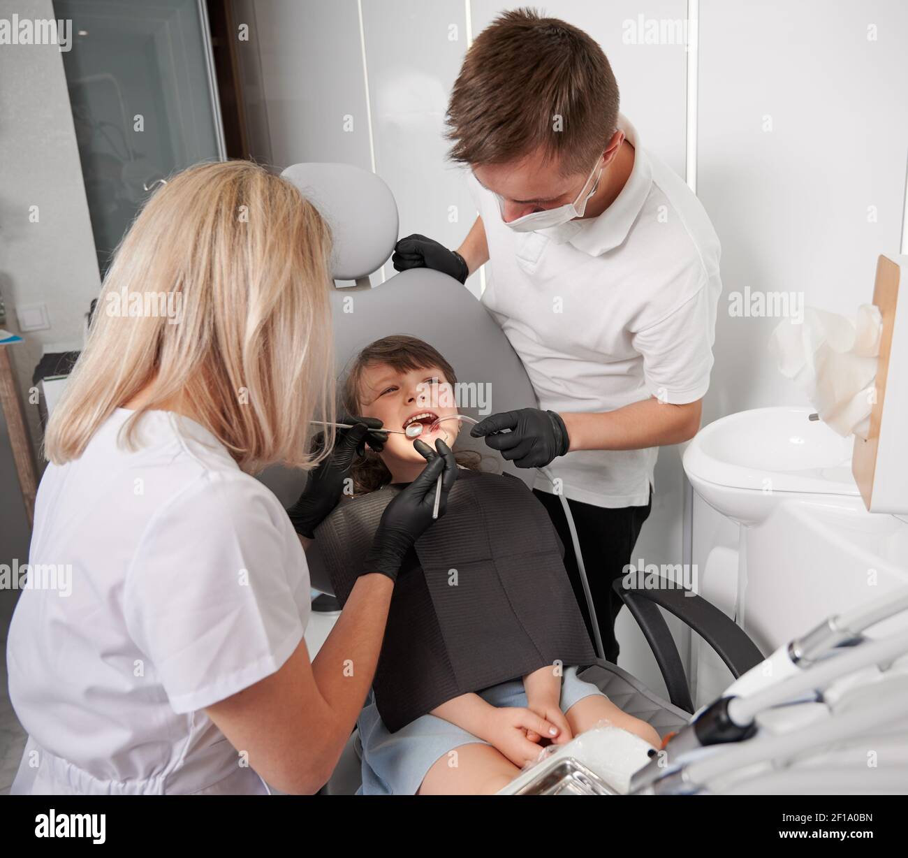 Bambino seduto in sedia dentale mentre due dentisti che controllano i denti del bambino. Dentista che esamina i denti della ragazza mentre l'assistente maschio tiene l'iniettore della saliva. Concetto di odontoiatria pediatrica e cura dentale. Foto Stock