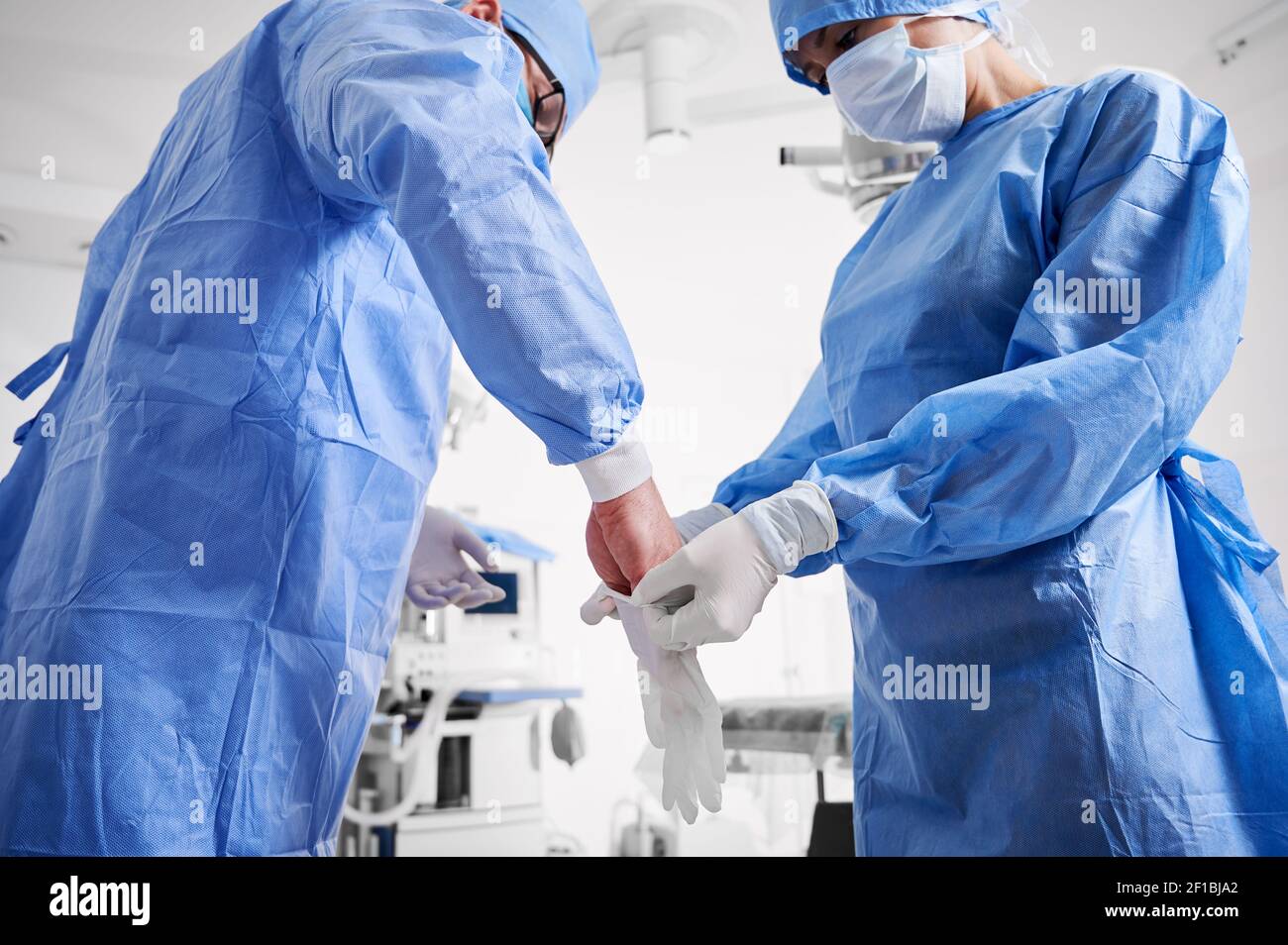 Assistente in tuta chirurgica blu che aiuta il chirurgo a indossare guanti  sterili. Due medici in maschere mediche protettive che si preparano per la  chirurgia plastica in clinica. Concetto di medicina e
