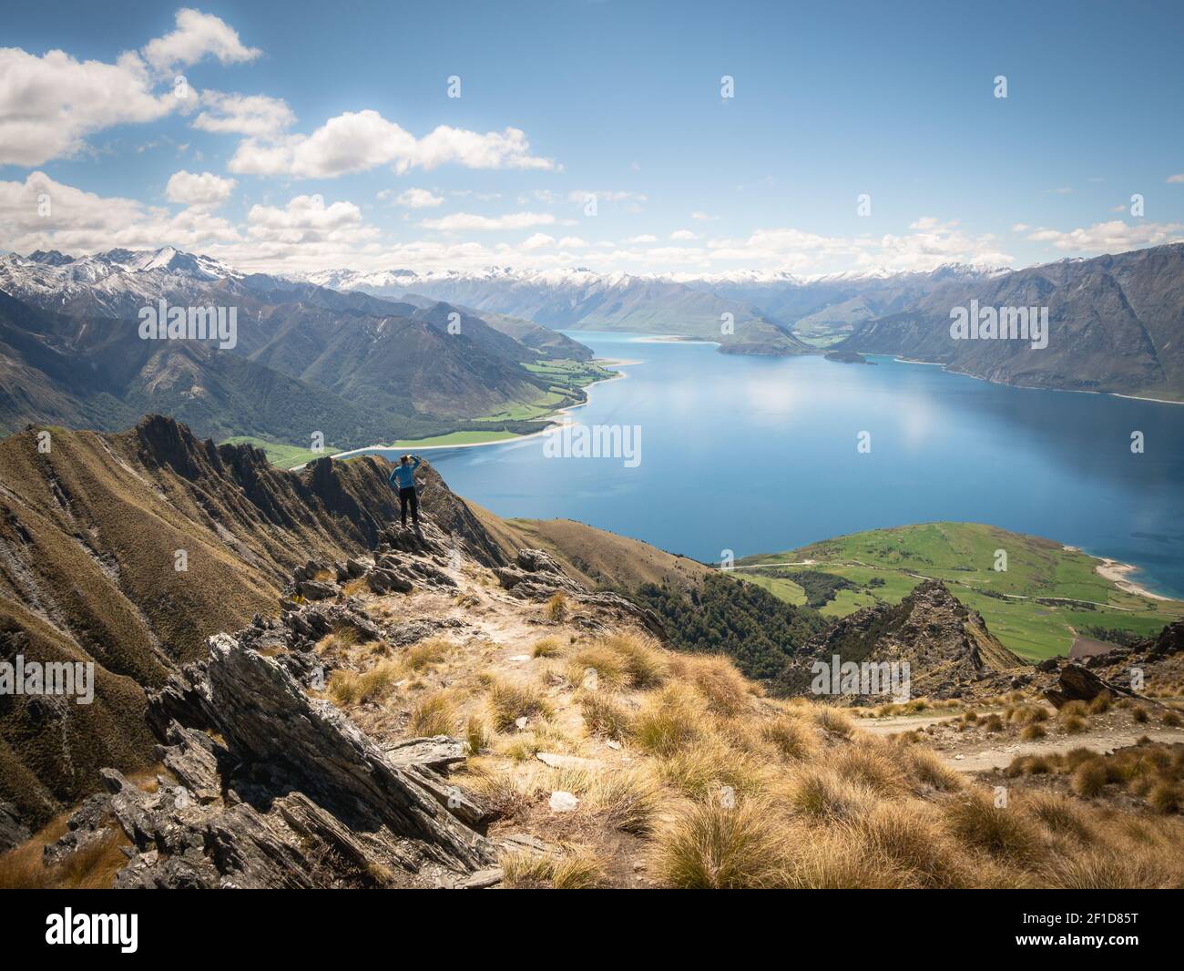Uomo in piedi sul bordo della roccia godendo di una splendida vista con prati verdi lussureggianti, lago blu e montagne innevate, giornata di sole sulla cima dell'Istmo Peak Foto Stock