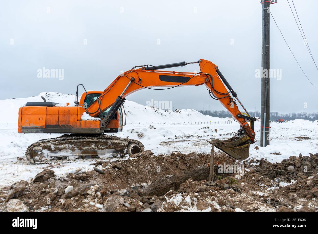 Un grande escavatore arancione scava il terreno per riparare e. posa di utenze e cavi sotterranei per l'elettricità e le comunicazioni Foto Stock