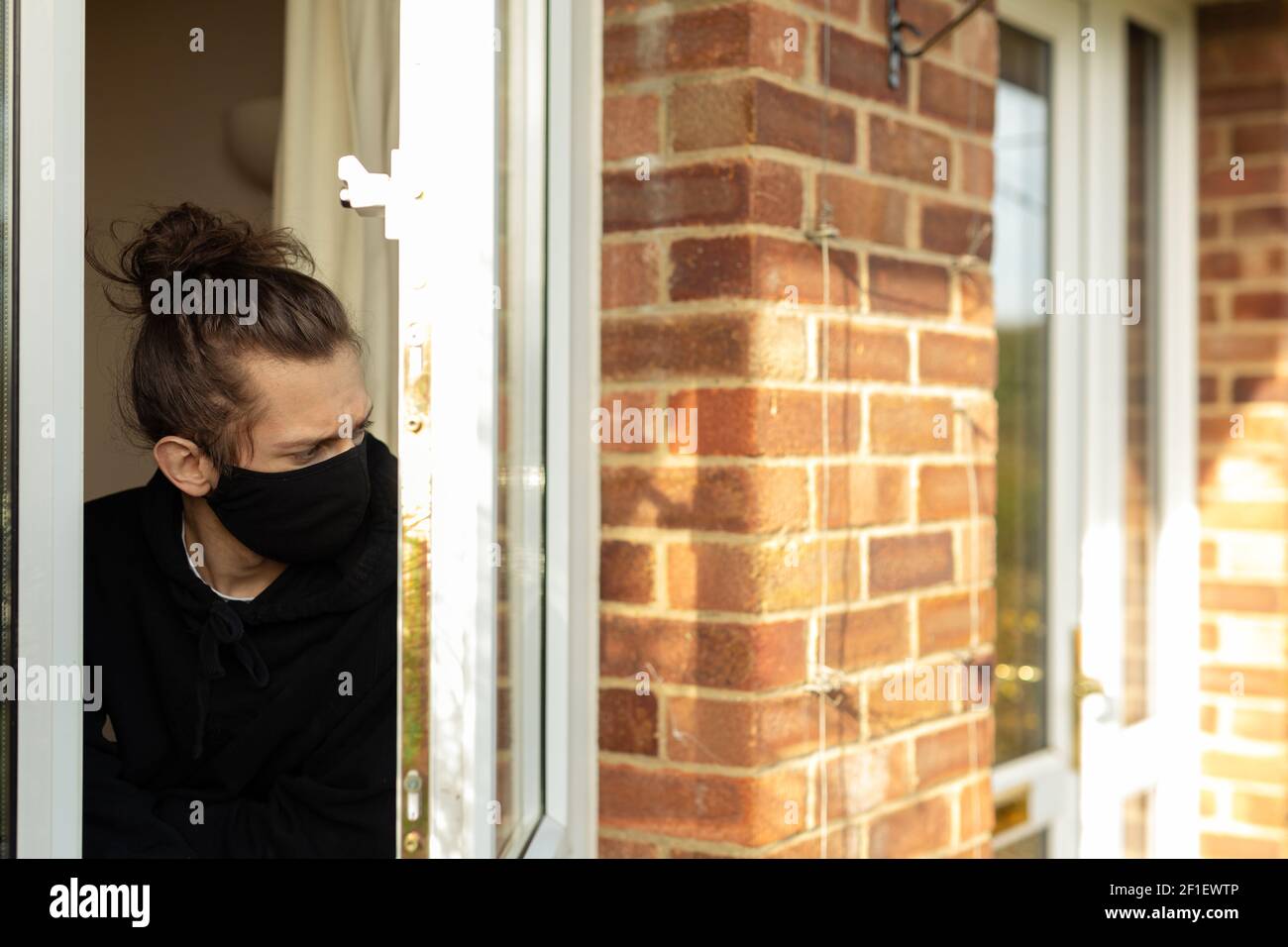 Un giovane uomo con una maschera facciale che guarda fuori la finestra durante il suo auto-isolamento a casa. Covid-19 concetto di solitudine di isolamento del virus corona Foto Stock