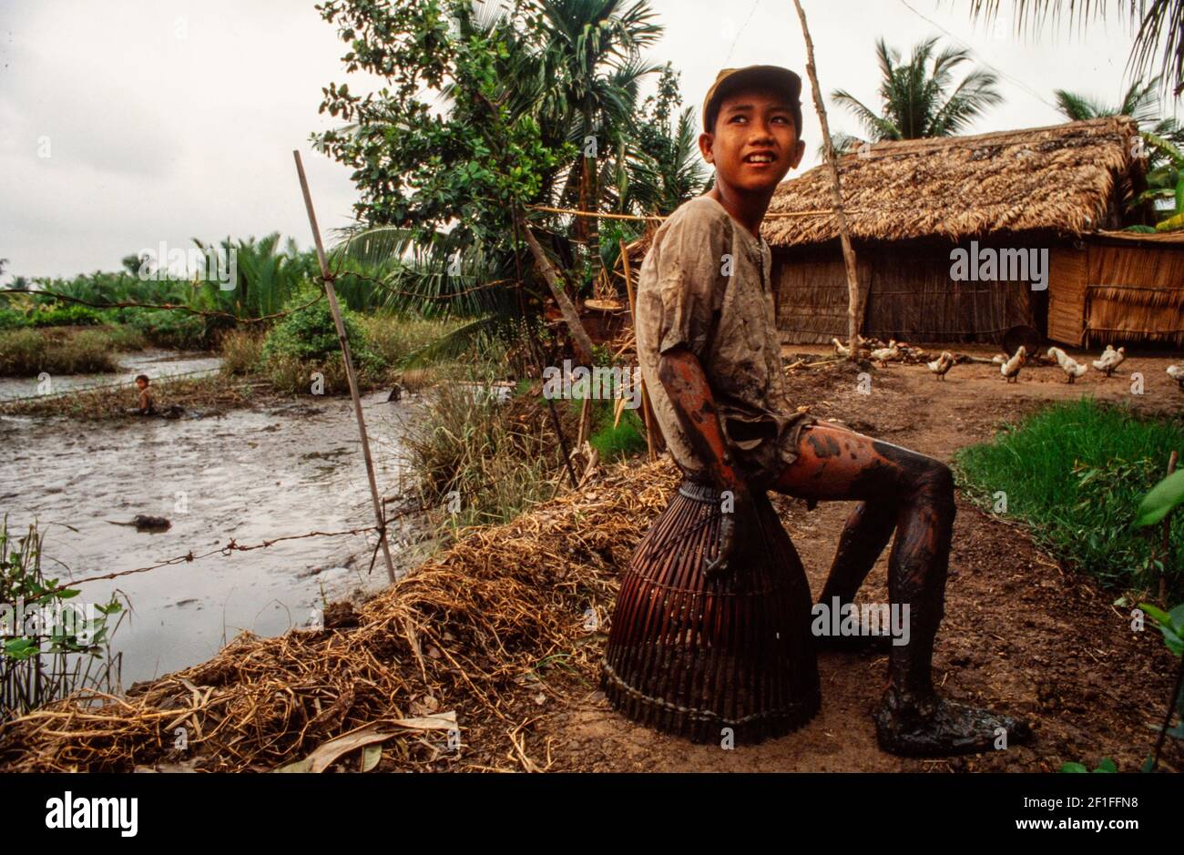 Un ragazzo si siede sul suo cesto di pesce dopo aver cacciato i granchi e il pesce nella risaia di famiglia, nel sud del Vietnam rurale, giugno 1980 Foto Stock