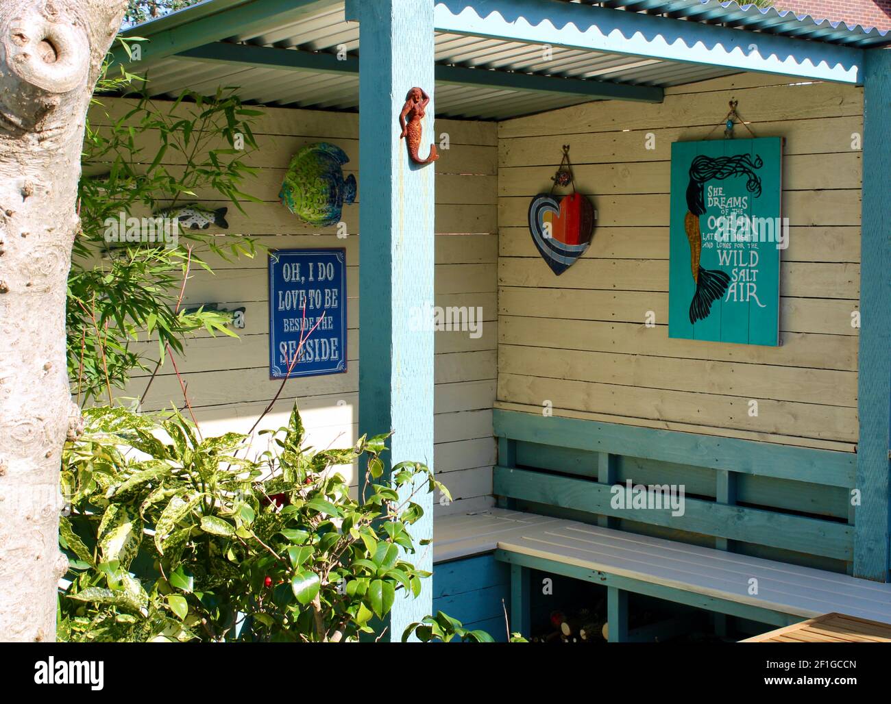 Area con posti a sedere nel giardino con decorazioni a parete che includono sirene, pesce, cuore e 'Io amo essere oltre il Poster Mare' Acuba japonica nelle vicinanze. Foto Stock