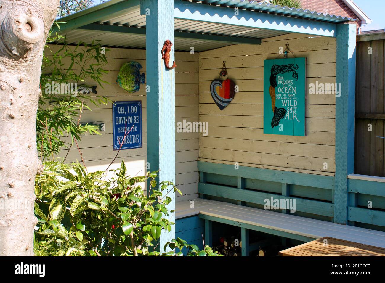 Area con posti a sedere nel giardino con decorazioni a parete che includono sirene, pesce, cuore e 'Io amo essere oltre il Poster Mare' Acuba japonica nelle vicinanze. Foto Stock