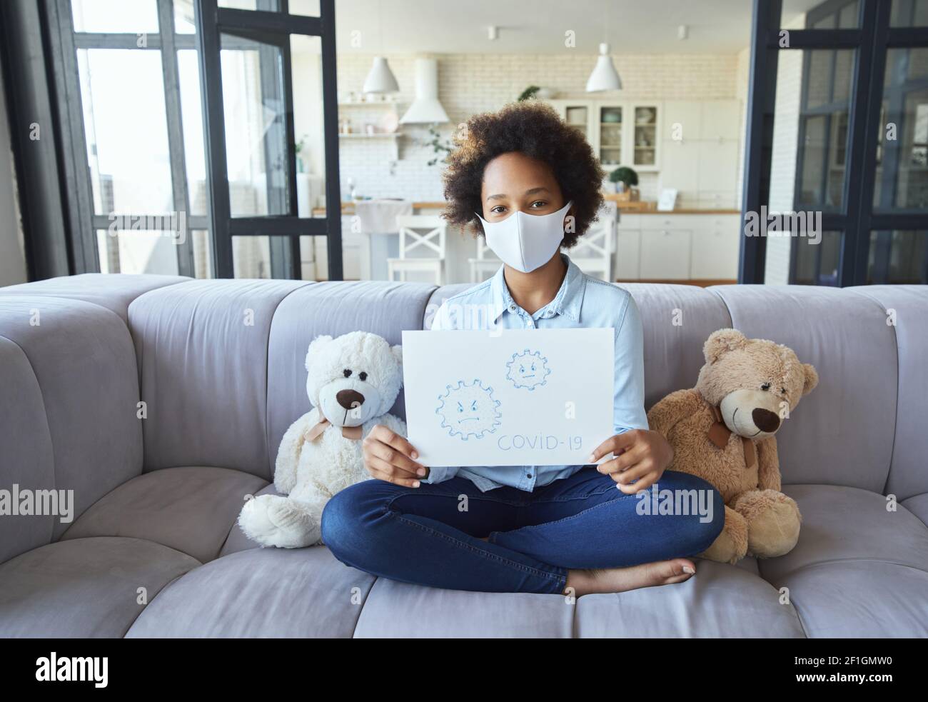 Misto razza teen ragazza indossare maschera protettiva guardando la macchina fotografica e tenendo Covid 19 disegno mentre si siede sul divano, studiando da casa durante Foto Stock