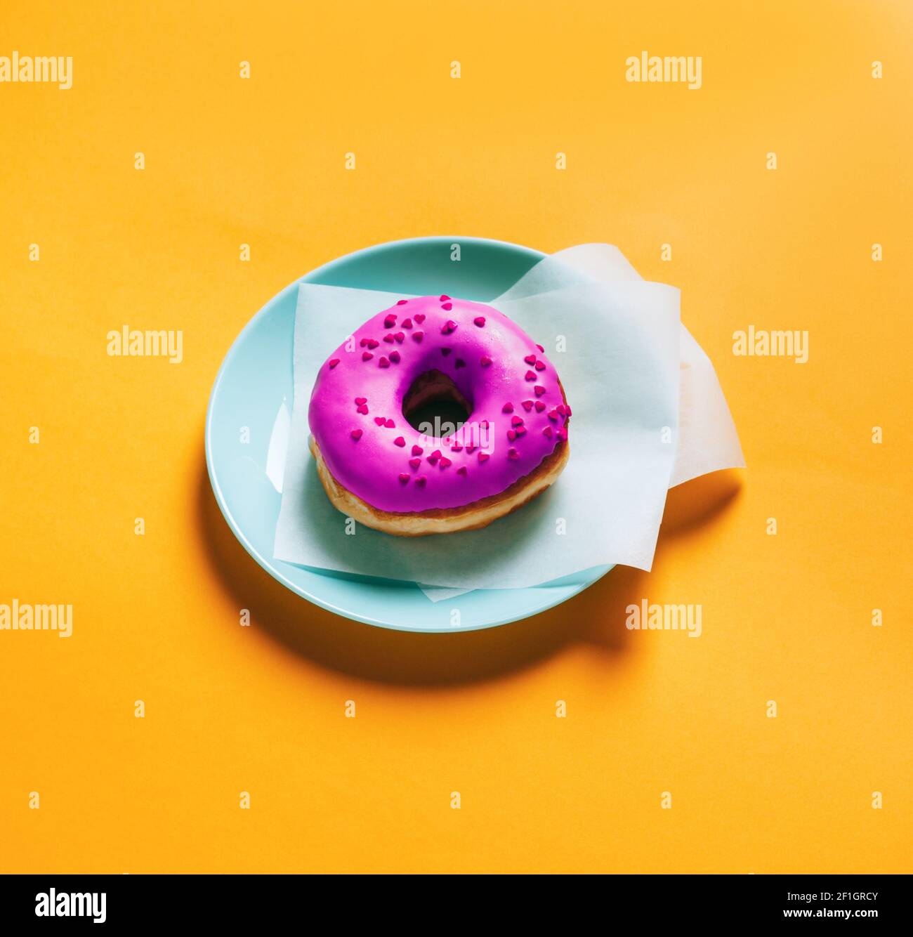 Sana ciambella porpora integrale con marmellata di frutti di bosco e cuore spruzzato su un piatto blu. Sfondo quadrato giallo isolato per la pubblicazione sui social media Foto Stock